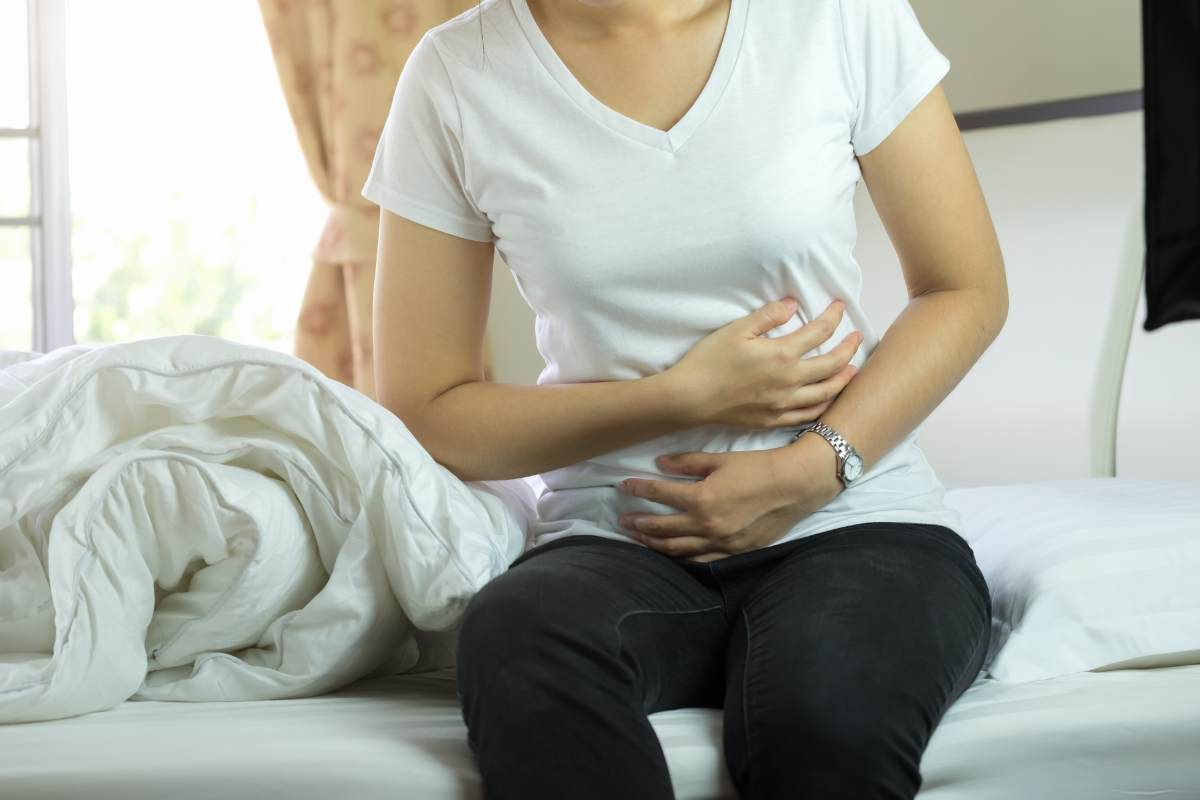 Mulher sentada na cama com a mão na barriga em expressão de dor abdominal