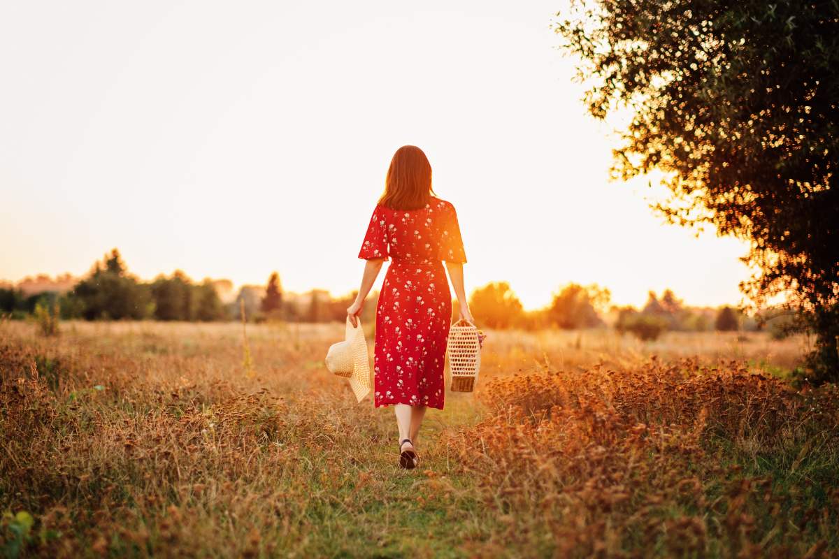 Mulher caminhando por um campo ao entardecer com chapéu e cesta nas mãos
